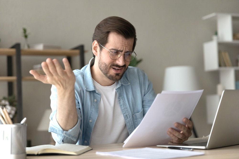 Заемщик в домашней или офисной обстановке с озадаченным видом просматривает бумаги, связанные с ипотекой или кредитным договором.