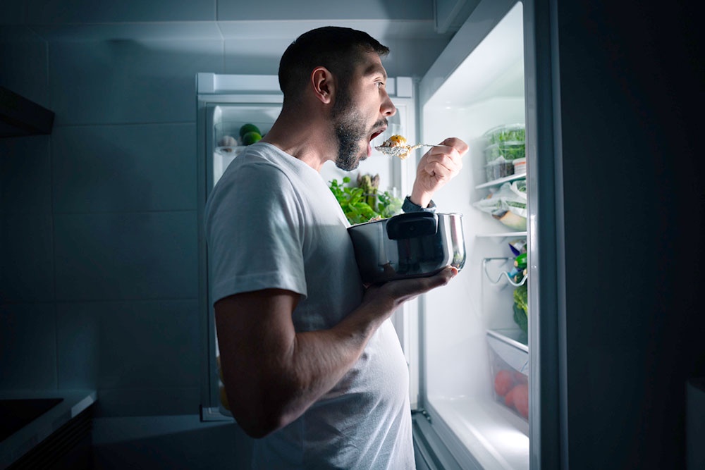 ночью мужчина есть салат из кастрюли возле открытого холодильника