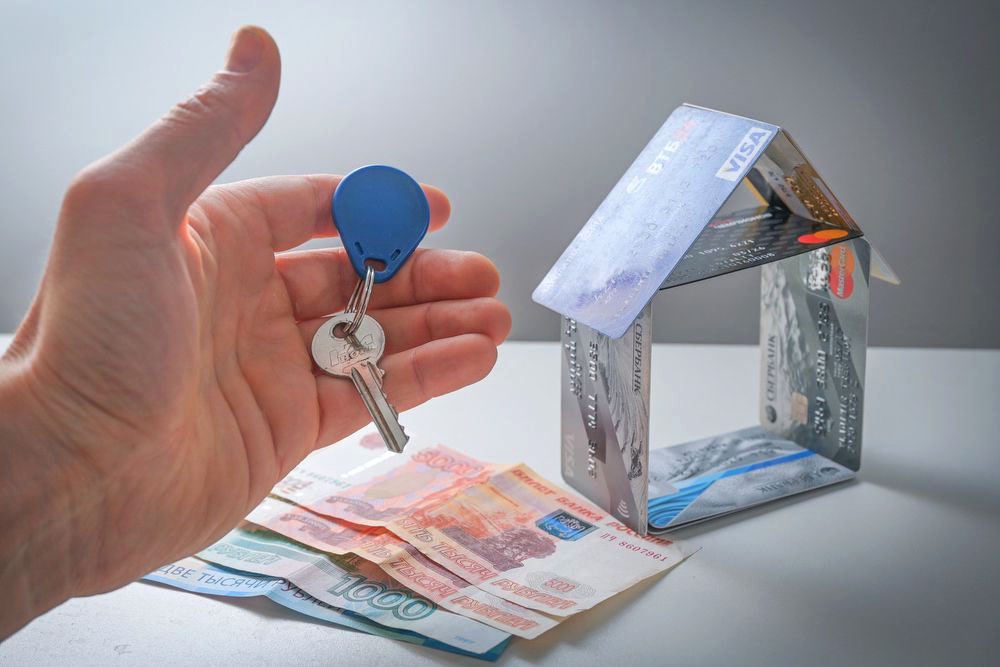 Человек держит ключи от квартиры на фоне денежных купюр и кредитных карт сложенных в виде домика