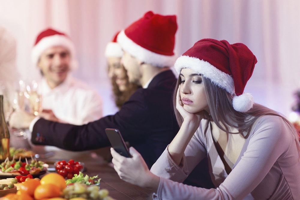 Грустная девушка сидит за праздничным и смотрит в телефон