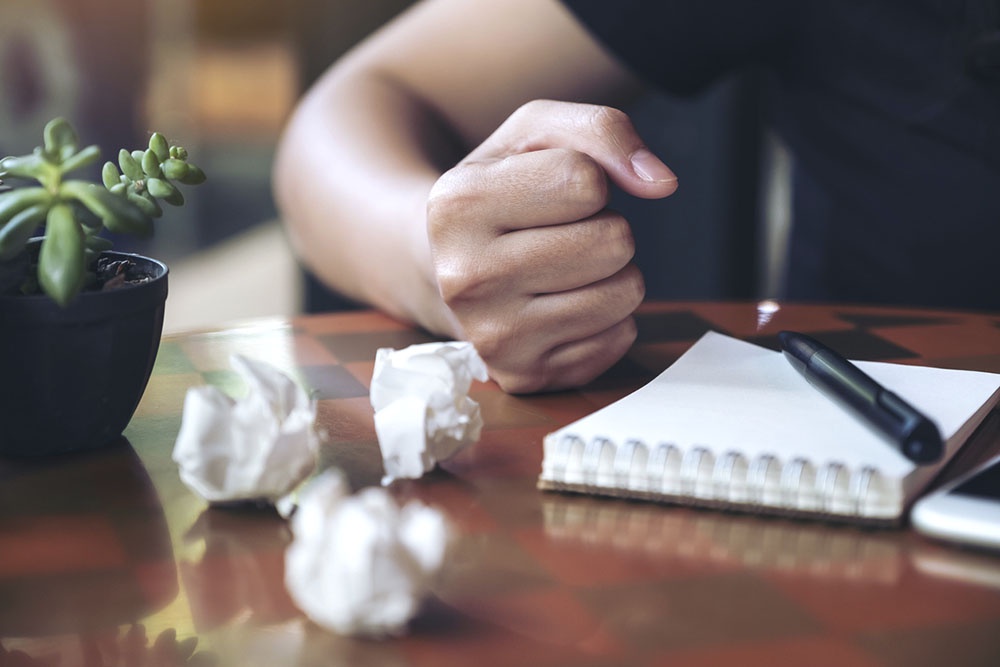 Человек сжимает пальцы в кулак и с негативными эмоциями пишет заметки в дневник