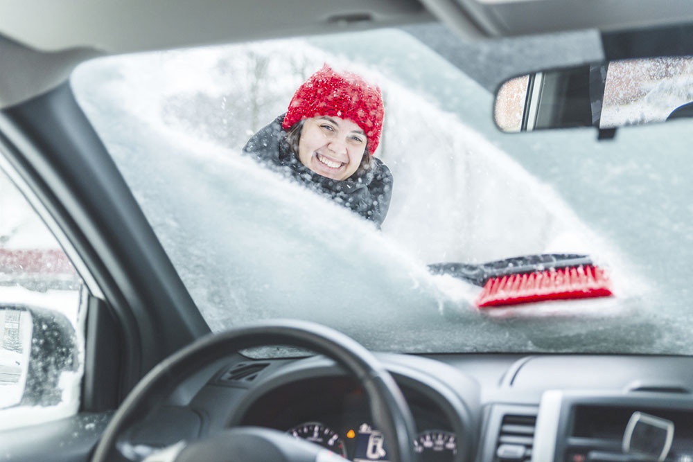 Девушка очищает лобовое стекло автомобиля от снега.