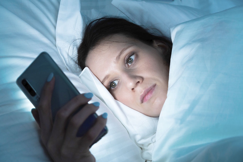 Женщина лежит в постели перед сном  и смотрит информацию на телефоне 