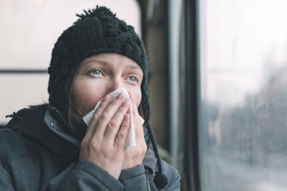 Девушка с насморком в общественном транспорте, вытирает нос платком сидя у окна.