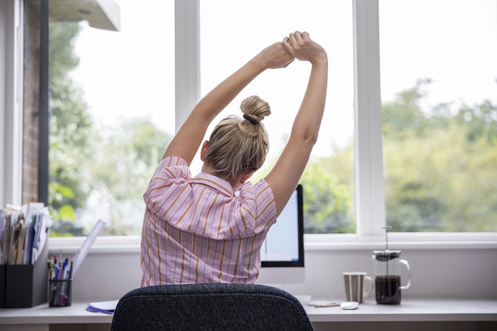 Женщина потягивается и разминает спину у рабочего стола, чтобы снять напряжение после нескольких часов за компьютером.