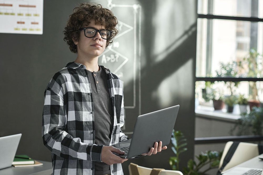 Школьник у доски в компьютерном классе объясняет код, написанный на ноутбуке.