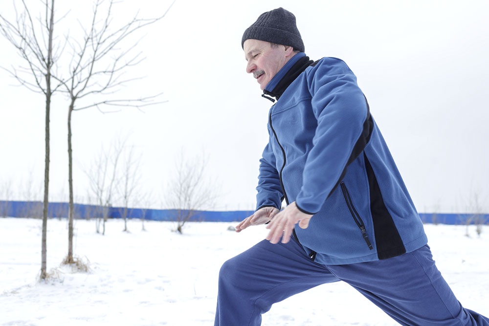 Мужчина выполняет легкую разминку с приседаниями перед уборкой снега на фоне заснеженного двора.