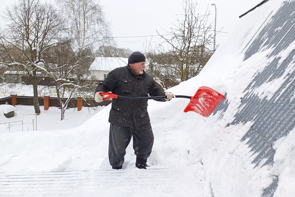 Мужчина очищает крышу частного дома от толстого слоя снега.