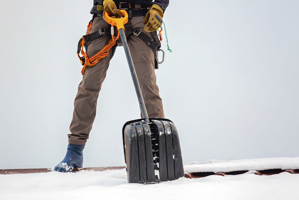 Работник в полной экипировке счищает снег стоя на нескользкой кровле.