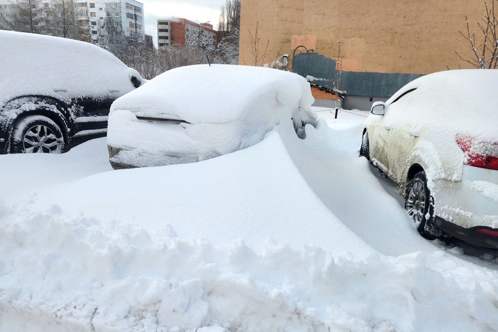 Несколько автомобилей, почти полностью засыпанных снегом, стоят на занесенной парковке или улице.