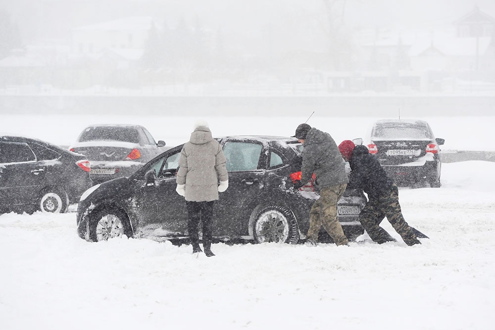 Несколько человек толкают застрявший в снегу автомобиль, водитель управляет рулем и газом.