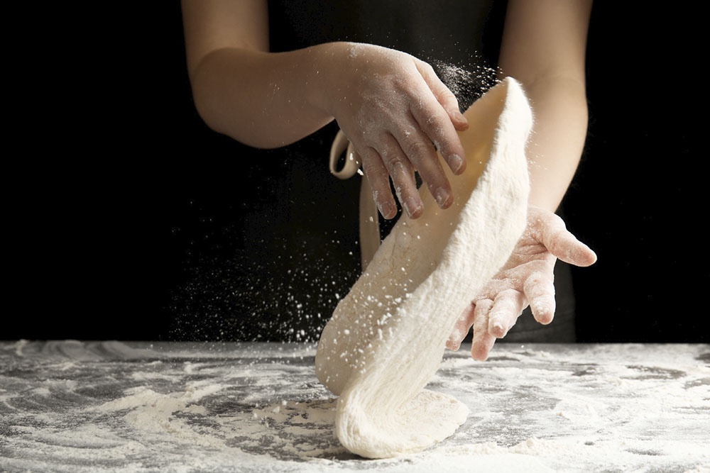 Крупный план рук, вымешивающих мягкое, эластичное тесто для пиццы на присыпанной мукой столешнице.