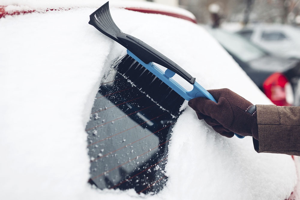 Водитель одной рукой счищает снег щеткой с лобового стекла.