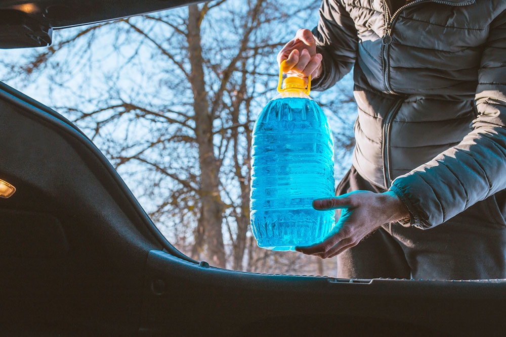 Водитель кладет канистру с незамерзающей жидкостью в багажник автомобиля на фоне зимнего пейзажа.