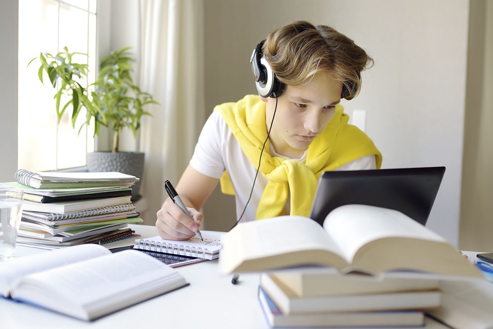 Школьник готовится к ЕГЭ по английскому дома: сидит за столом с учебниками, тетрадями и ноутбуком.