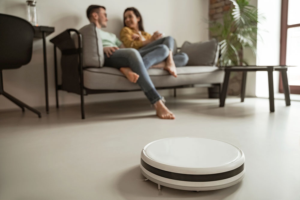 Мужчина и женщина пьют чай на диване, пока робот-пылесос на переднем плане автоматически убирает пол в комнате.