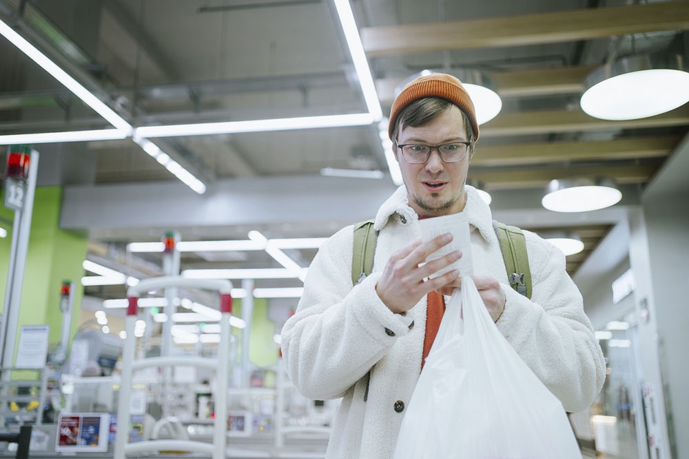 Мужчина изучает чек в магазине и огорчен итоговой суммой покупок.