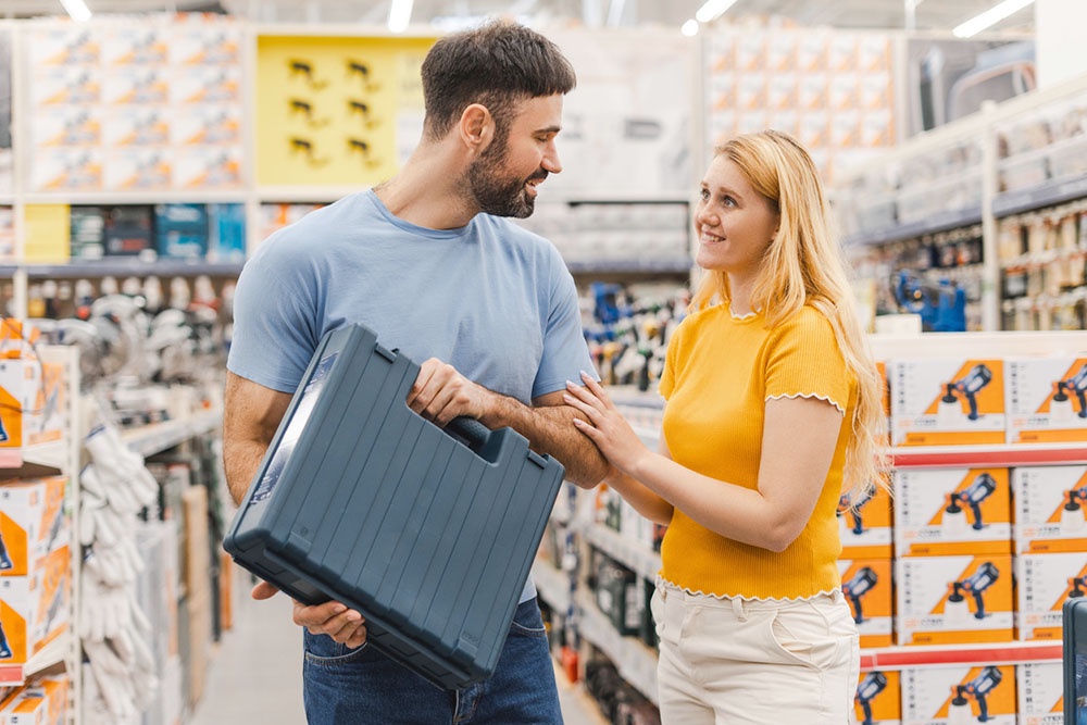 Пара в строительном магазине: женщина помогает мужчине выбрать органайзеры и боксы для инструментов. 