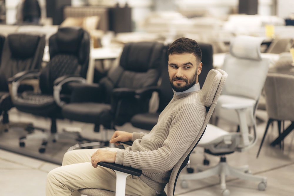 Мужчина выбирает качественное компьютерное кресло для дома, примеряясь к разным моделям и оценивая удобство.