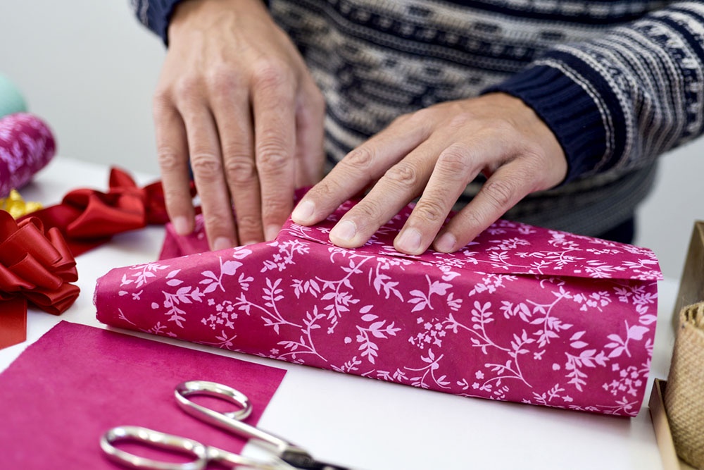Мужчина упаковывает подарок в красивую оберточную бумагу, старательно разглаживая складки и завязывая ленту.