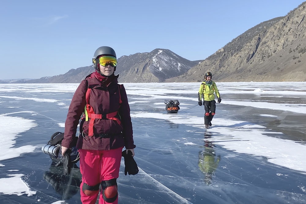 Туристы идут в поход по замерзшему озеру Байкал зимой. Группа путешественников в теплой одежде с рюкзаками пересекает прозрачный лед на фоне заснеженных берегов острова.