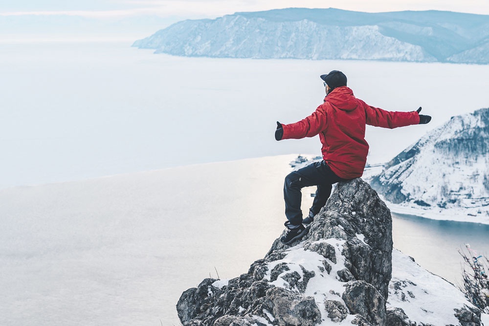 Турист сидит на Черском камне и любуется живописным видом на озеро Байкал и исток реки Ангары в поселке Листвянка. Один из самых известных смотровых пейзажей на Байкале, открывающий панораму замерзшего озера и незамерзающего ангарского течения.