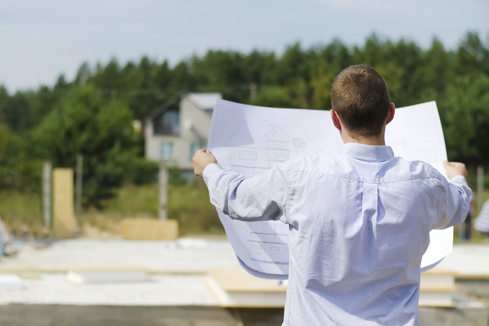 Человек осматривает участок и проект дома на месте строительства.