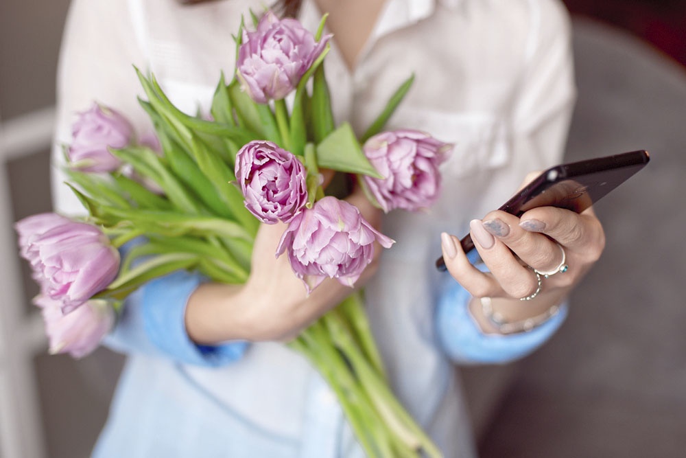 Счастливая женщина с букетом тюльпанов читает поздравительное сообщение с 8 Марта в телефоне.