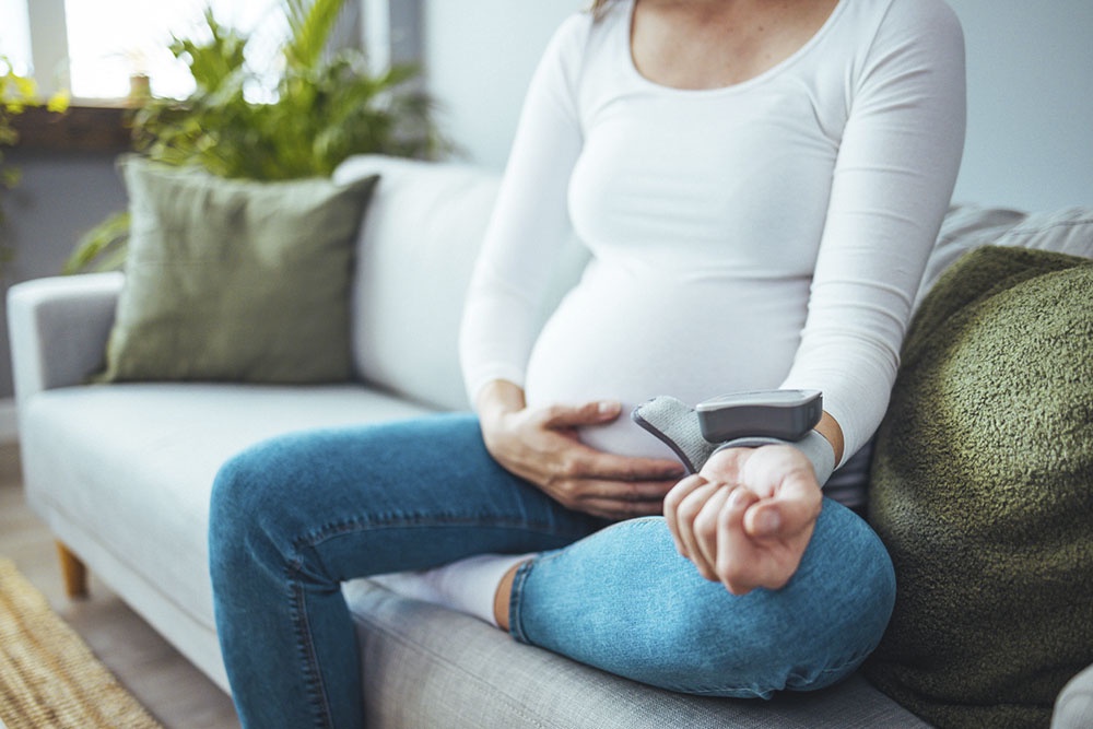 Беременная измеряет давление дома сидя на диване.