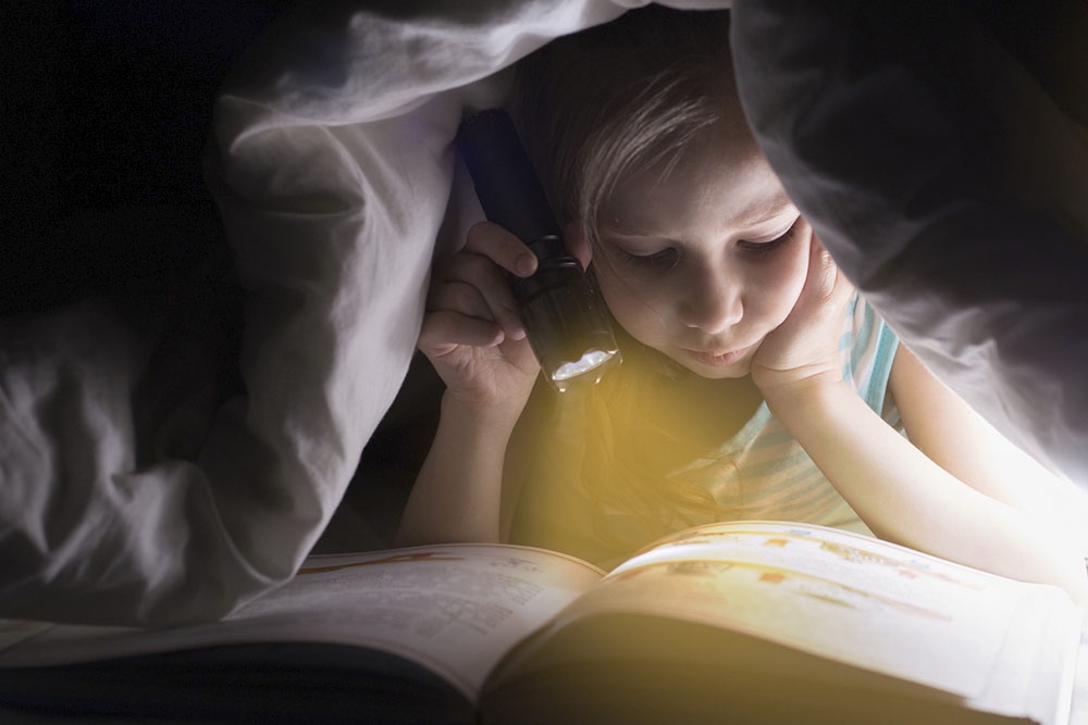 Ребенок читает книгу ночью под одеялом.