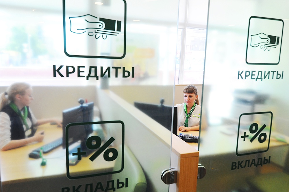 Сотрудницы банка сидят за стеклянными дверьми, на которых написана информация о вкладах, кредитах, ипотеке