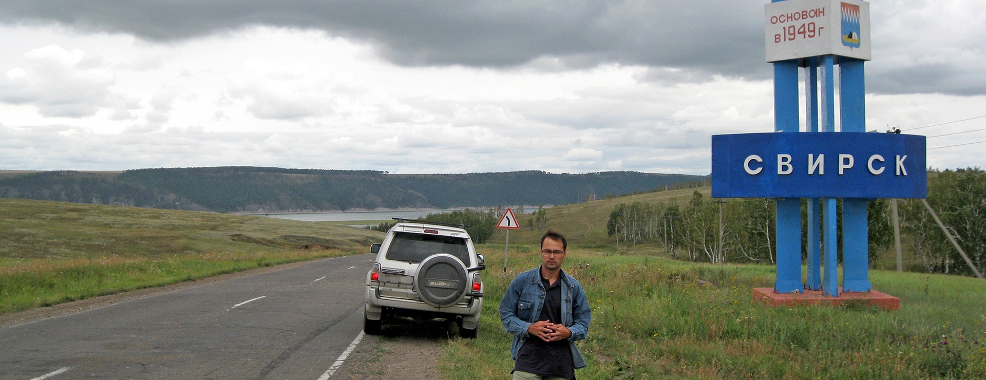 Авто на обочине асфальтированной дороги, справа стела с надписью "Свирск". На переднем плане Василий Авченко