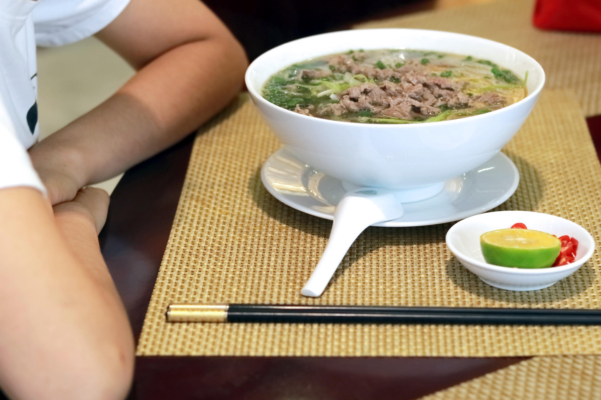 Миска с супом фо бо, ложка, палочки. Человек сидит за столом.