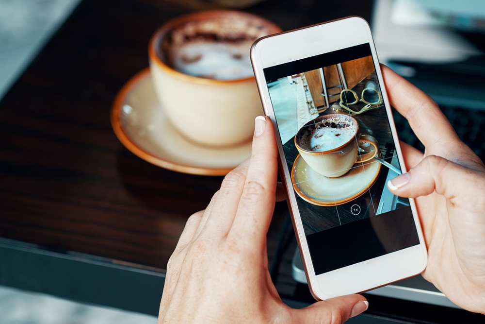 Женщина фотографирует чашку кофе при помощи смартфона.