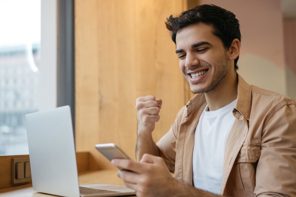 Вероятны плодотворные деловые переговоры, возможно начало сотрудничества на новых условиях. Мужчина перед ноутбуком радуется, держа телефон в руке.