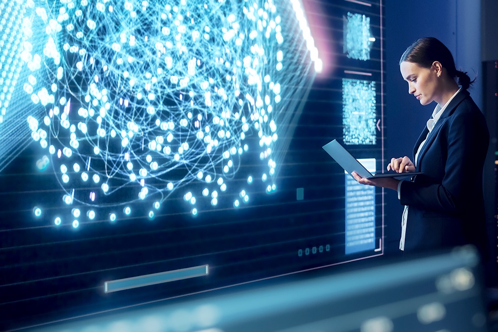 Девушка с ноутбуком стоит возле большого экрана.