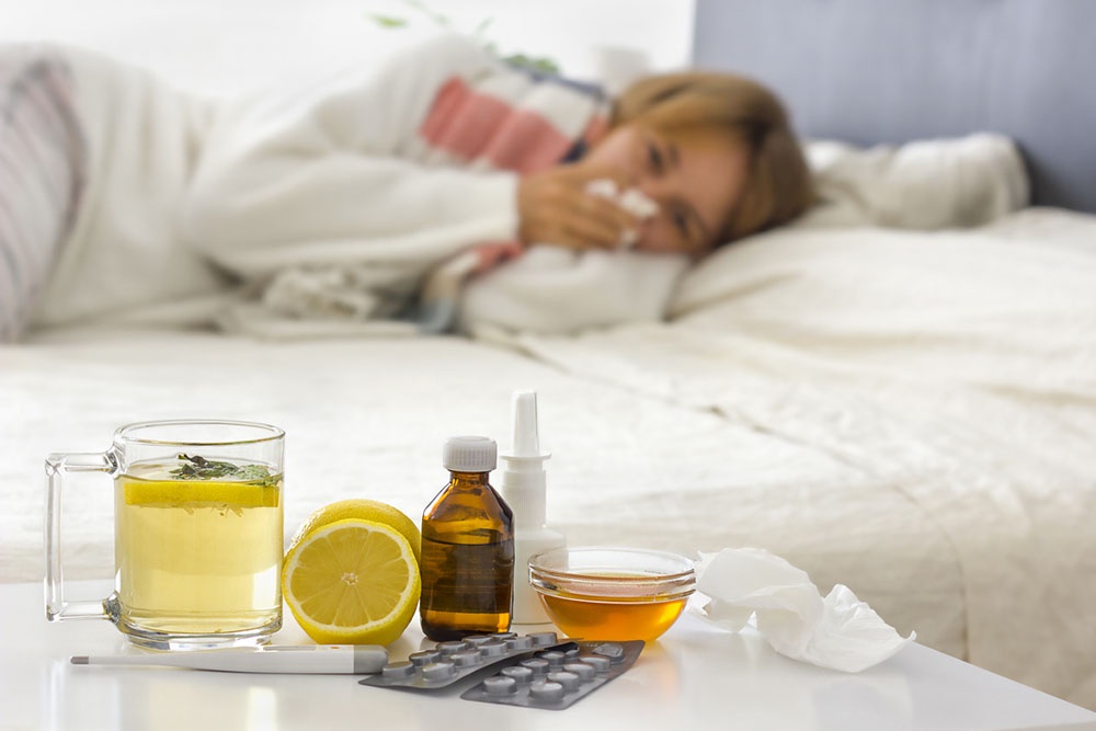 Больная девушка в постели, на тумбочке рядом стоят чашка с чаем и упаковка таблеток.